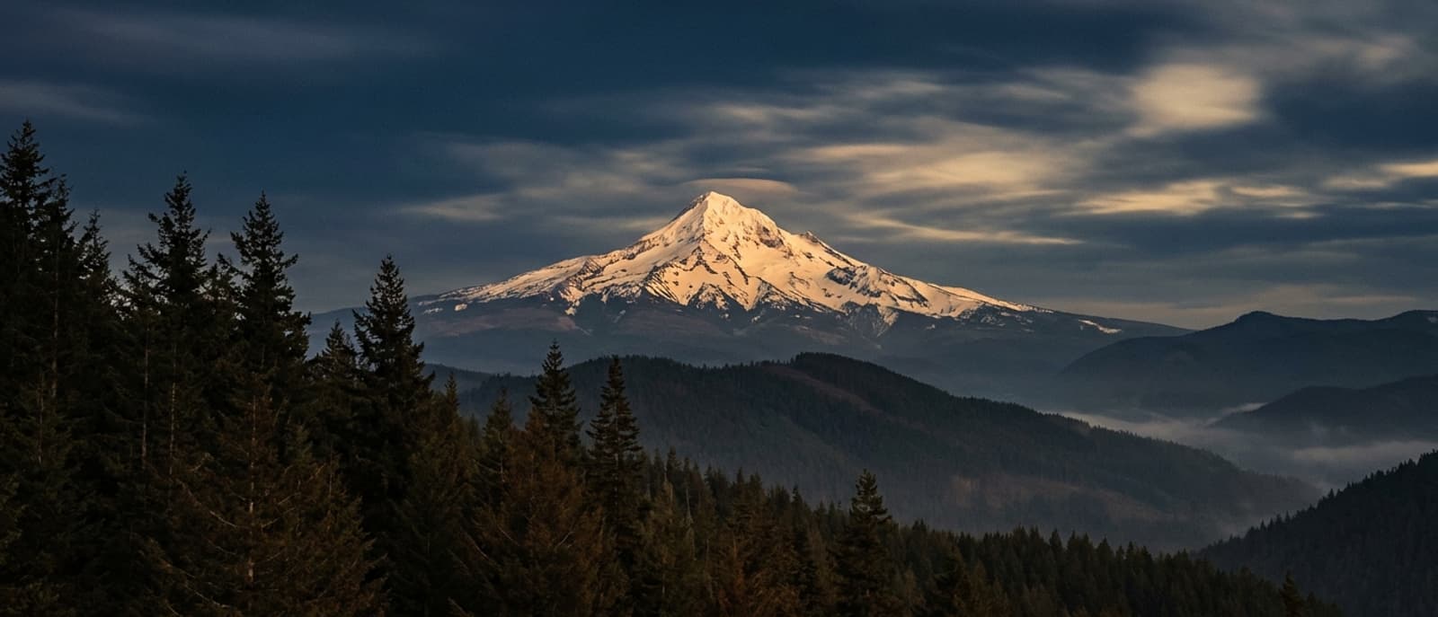Mount Hood at dusk above dark evergreen ridgelines, Oregon